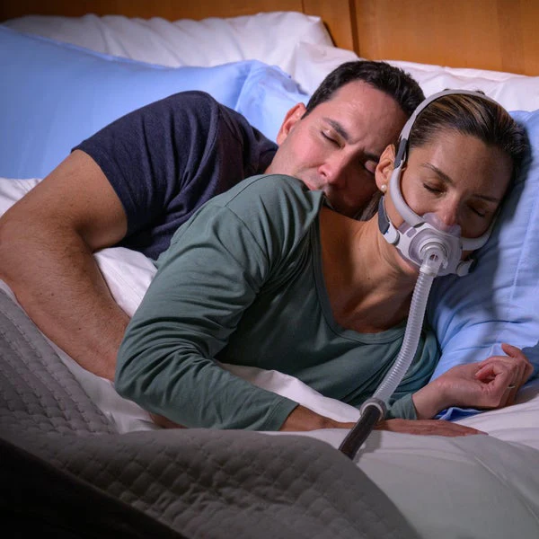 AirFit40_AirCurve11_Couple-Bed-Sleeping_3796_720x Kobieta śpi w łóżku z aparatem CPAP na twarzy, obok niej leży mężczyzna.