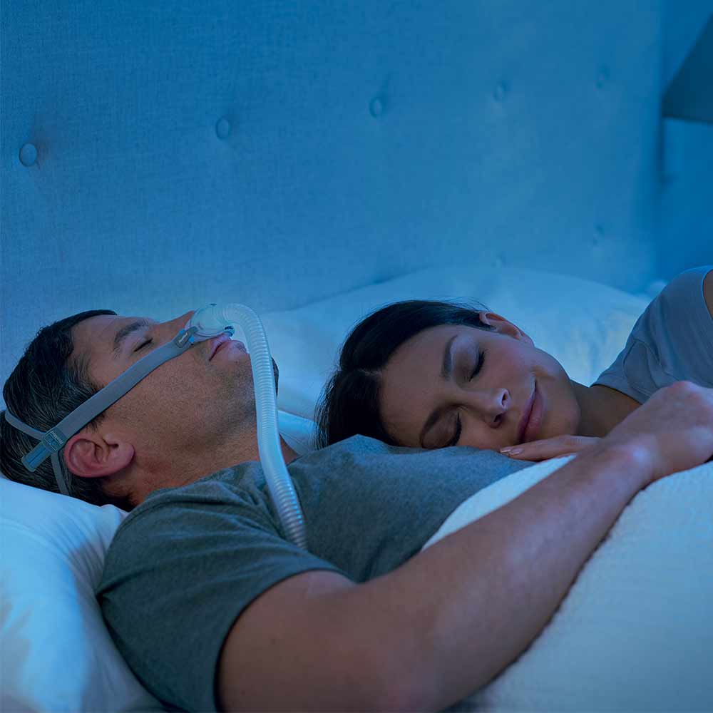 maska-donosowa-airfit-p10-sklep-resmed Mężczyzna śpi z aparatem CPAP, kobieta leży obok niego, oboje odpoczywają w łóżku.