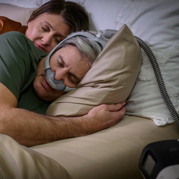 AirTouchN30i_Couple-Sleeping_0720_720x Para śpi w łóżku, mężczyzna używa maski CPAP do leczenia bezdechu sennego.
