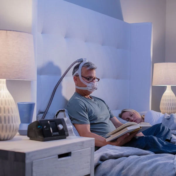 F30i-1E1A4626-Edit_720x Mężczyzna z maską CPAP czyta książkę w łóżku, kobieta obok niego śpi.