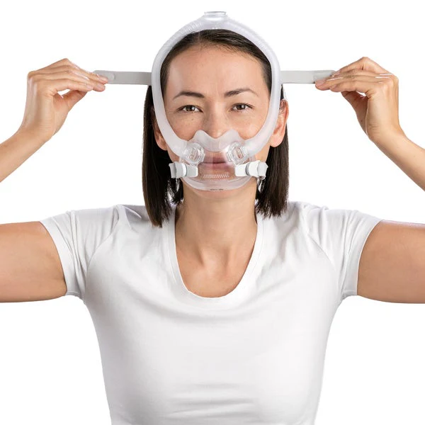 F30i_Fitting_Female_4_720x Kobieta zakłada maskę CPAP na twarz, trzymając paski po bokach głowy.